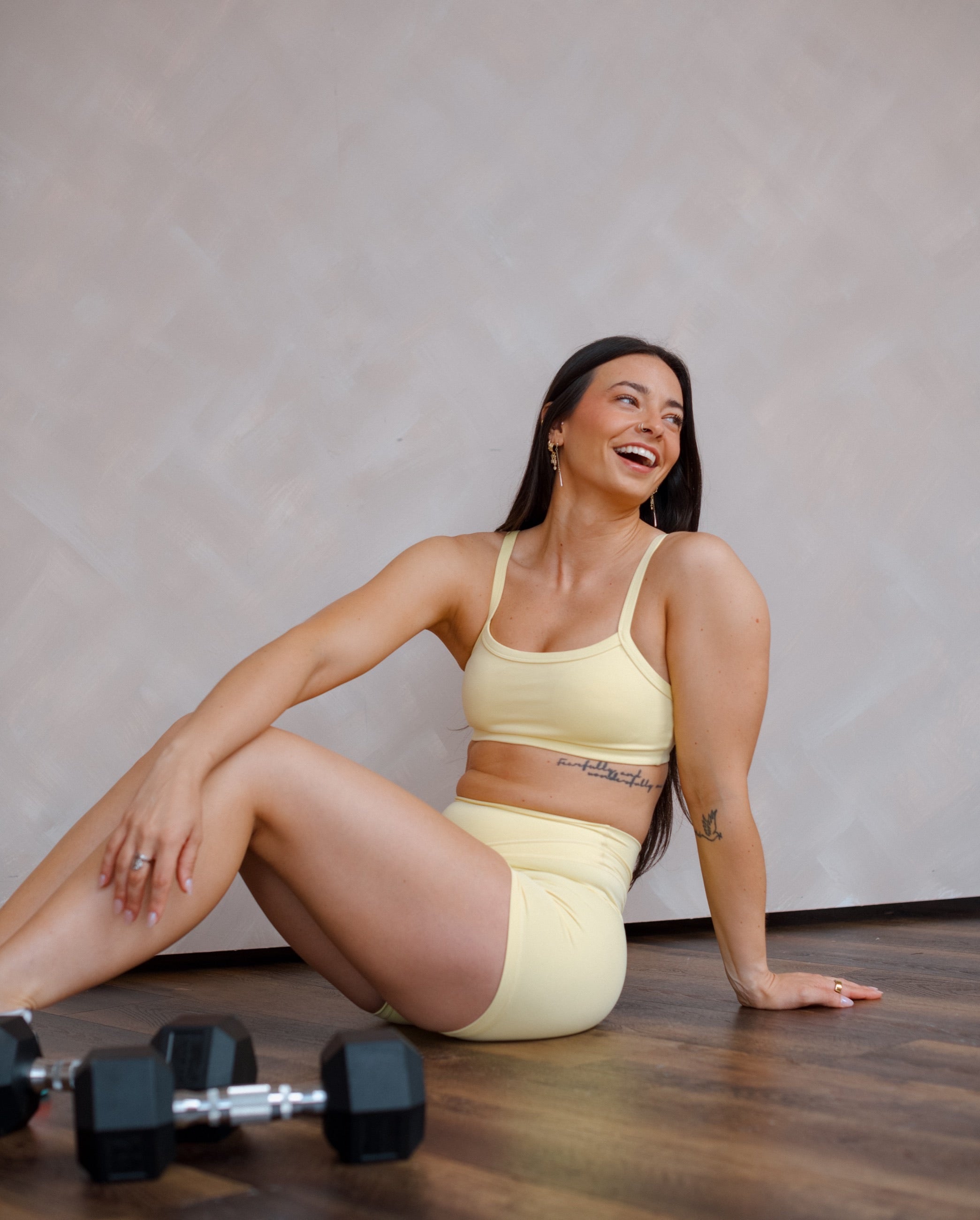 A woman laughing, seated on the floor in yellow activewear beside a dumbbell